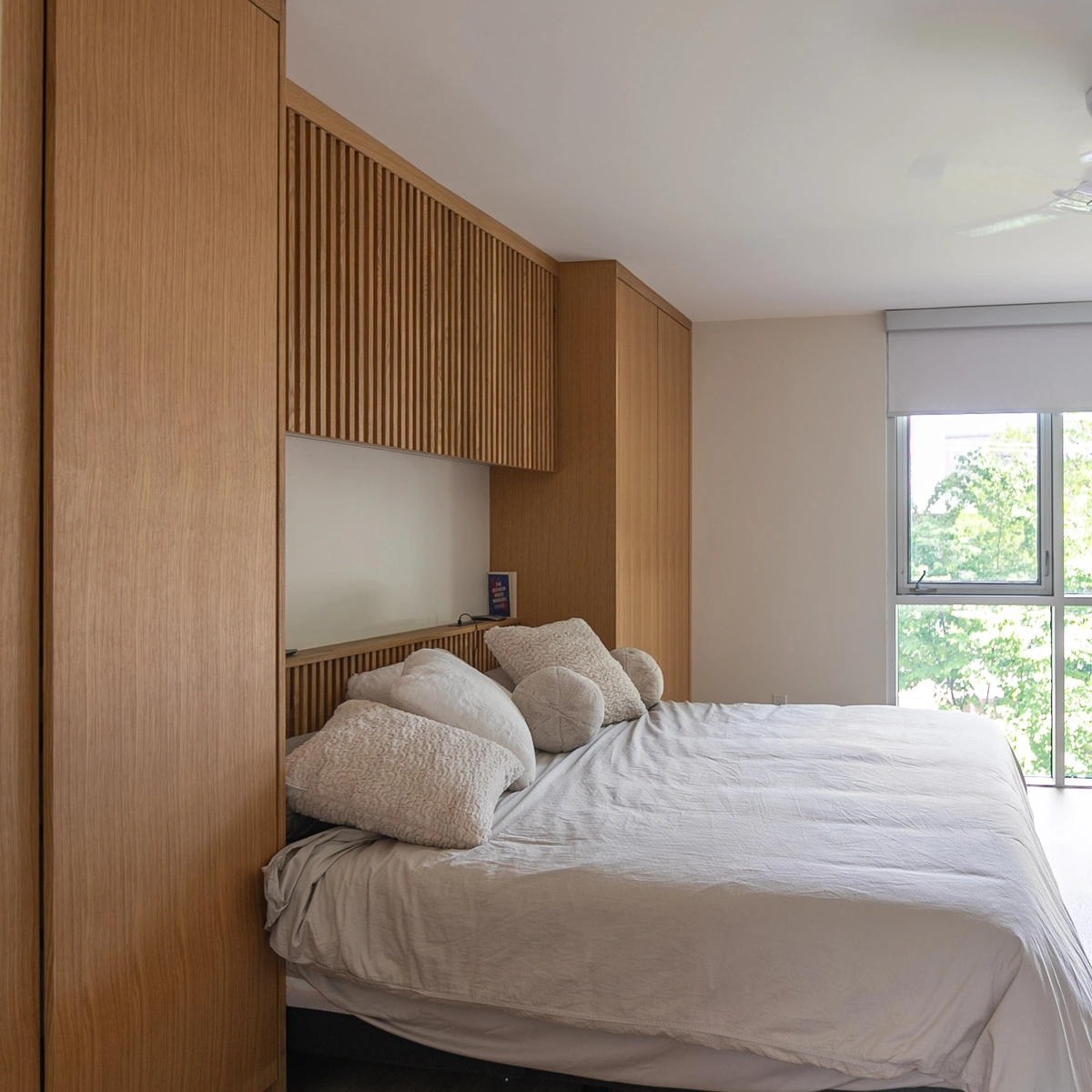 Custom overbed wardrobe in light oak framing a bed with slatted upper cabinets